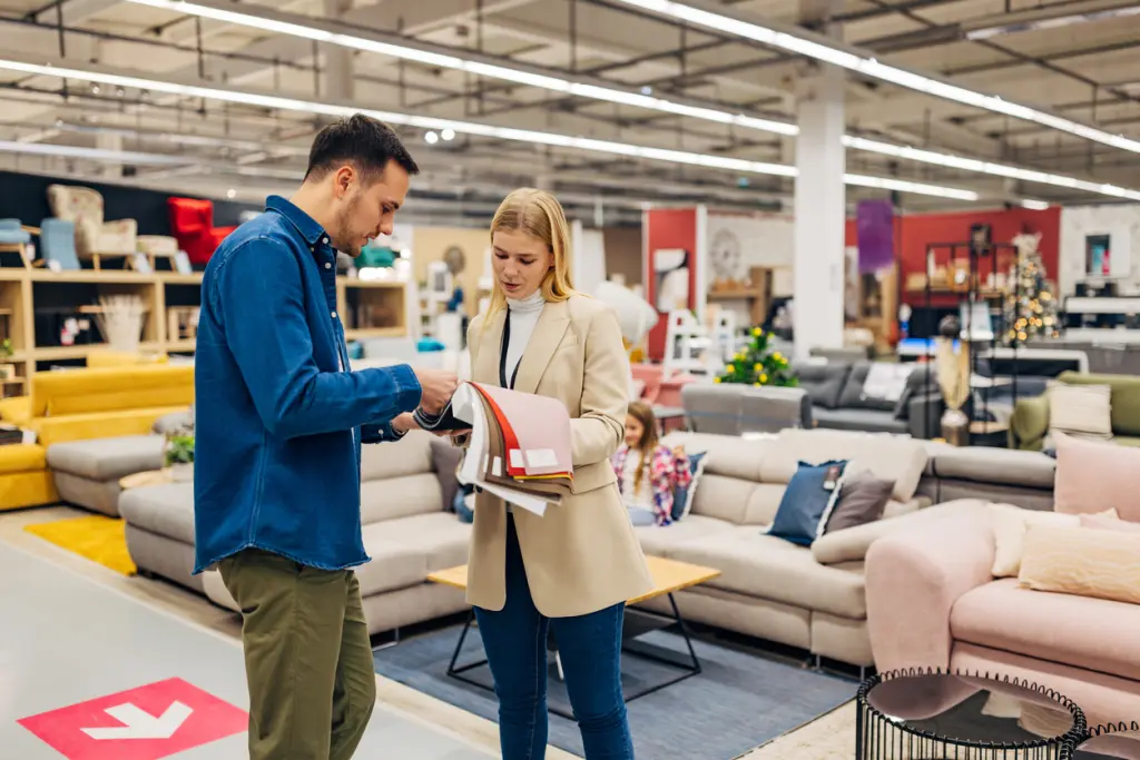 Sales associate showing fabric swatches to a customer in a modern furniture store, with sofas, coffee tables, and home decor visible in the background.